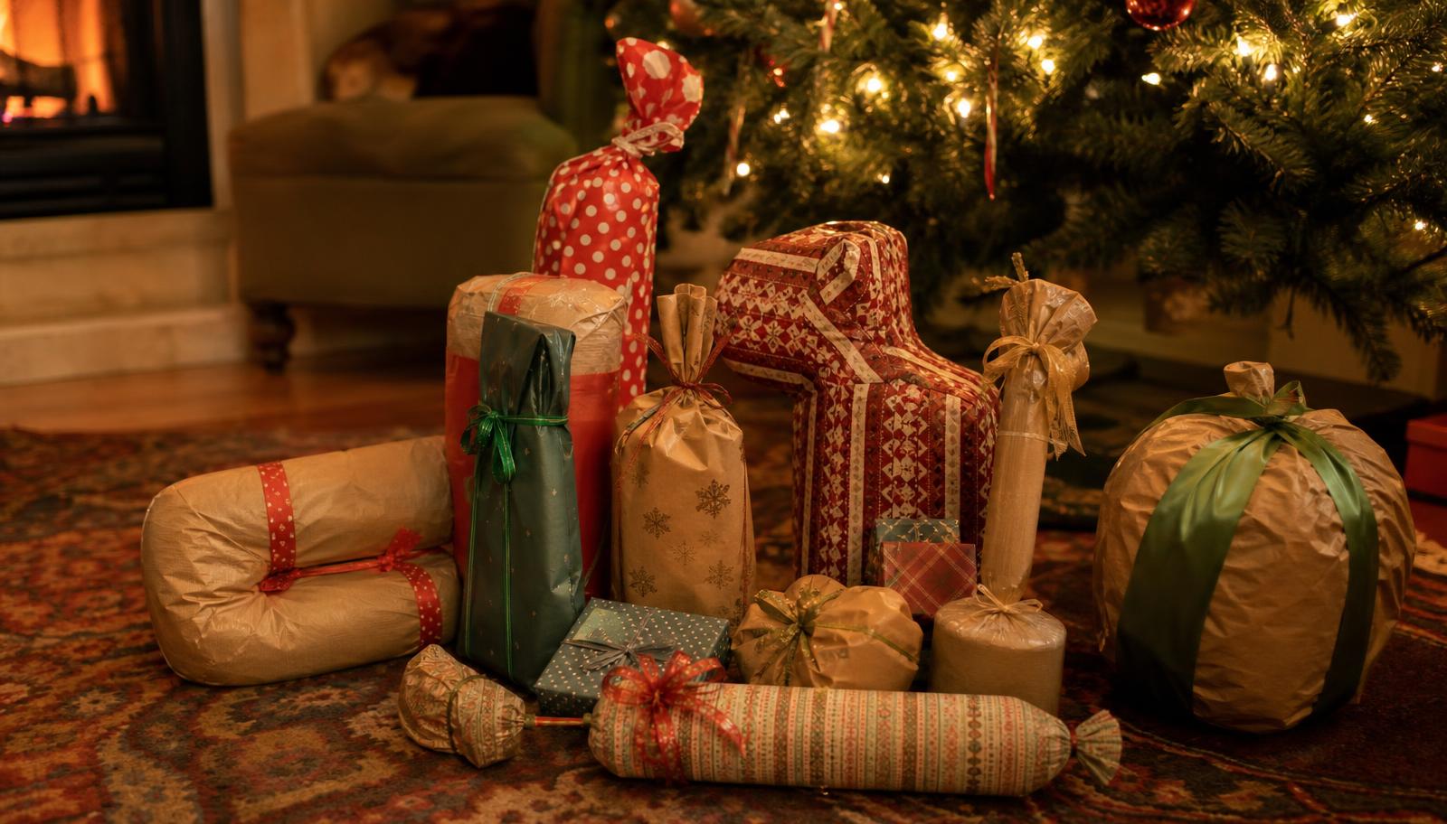 A pile of oddly shaped gifts wrapped in mismatched paper stacked on a rug beside a lit Christmas tree and a fireplace